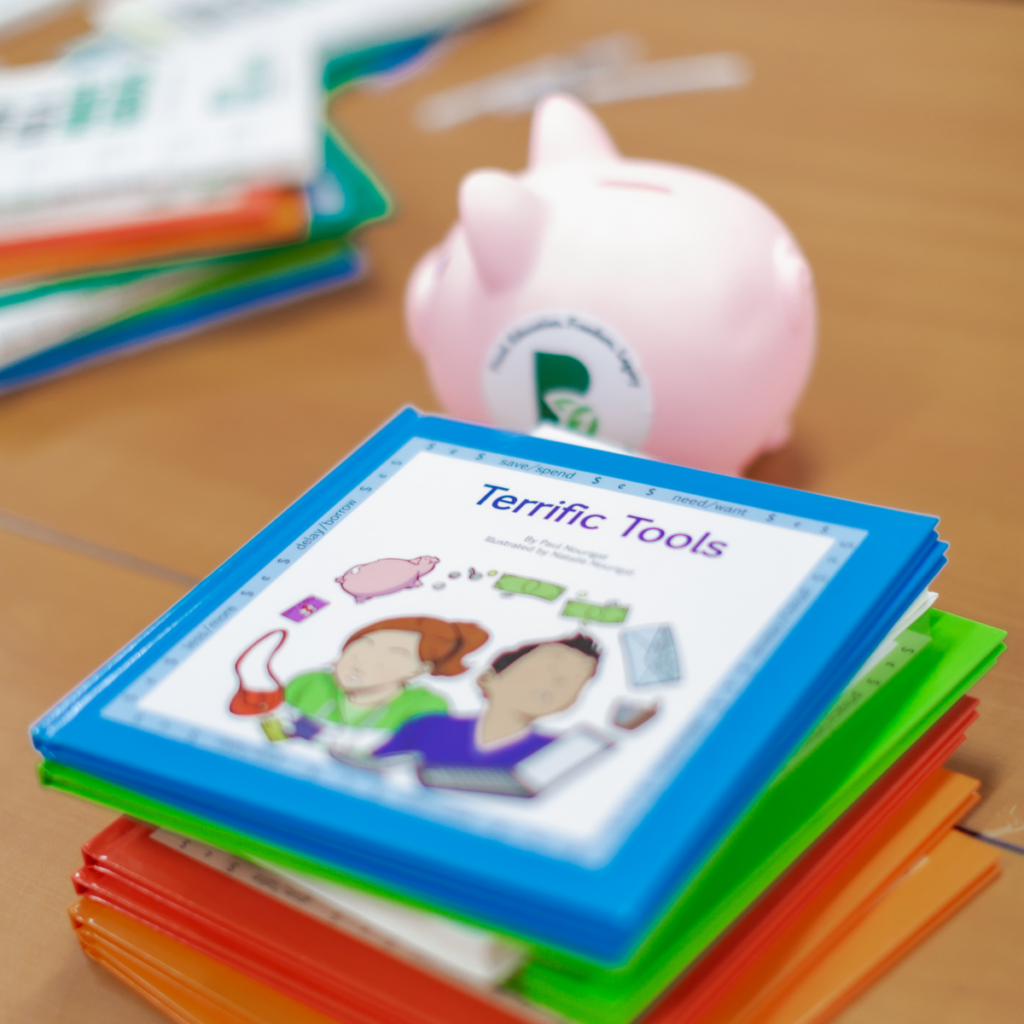 stack of childrens financial books: blue, green, red, and orange borders with top book reading "Terrific Tools". Books stacked in front of piggy bank.